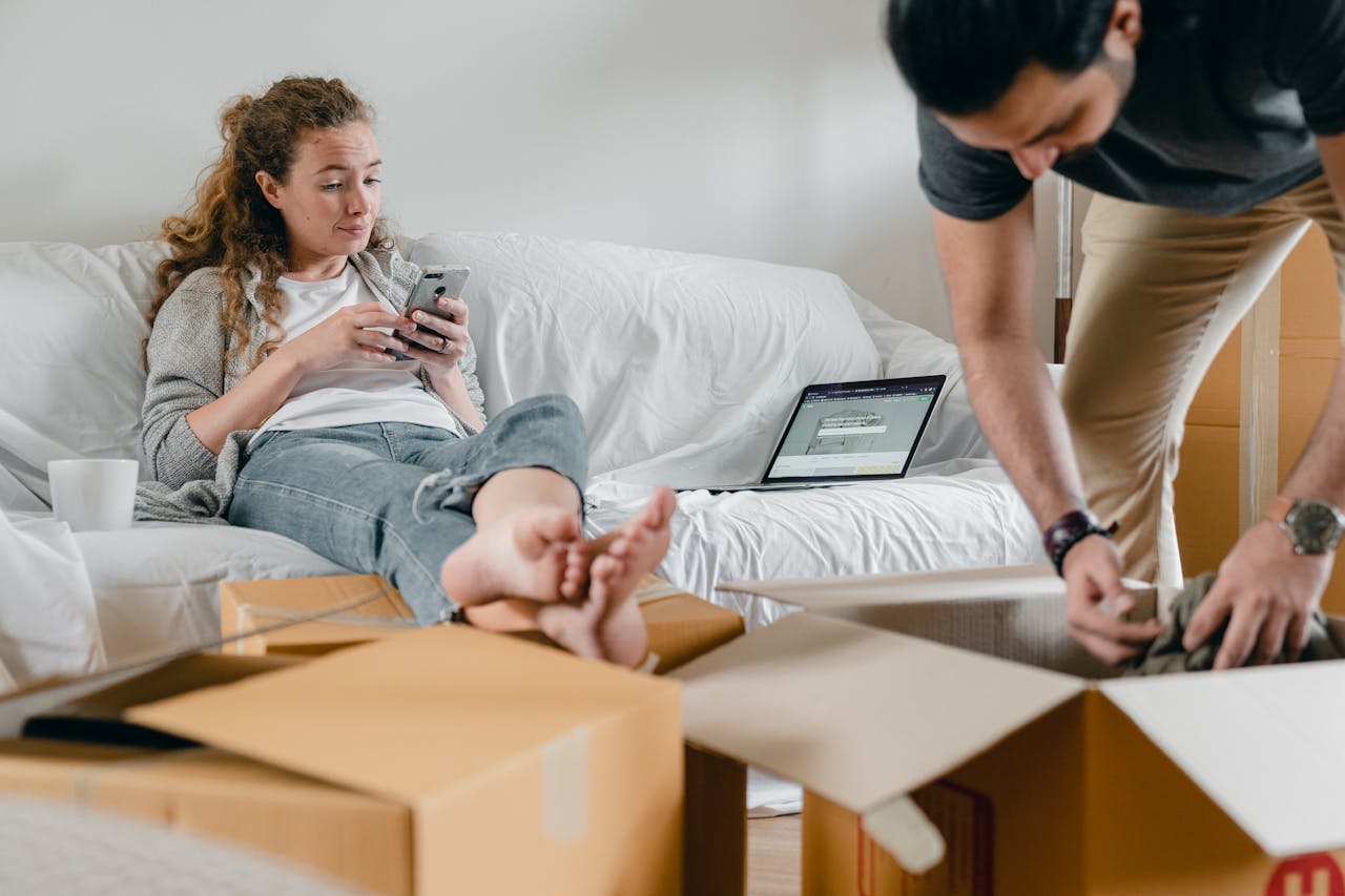 Barefoot female in casual wear sitting with crossed legs on sofa with open laptop while using social media on cellphone and crop male partner getting clothes out of cardboard box